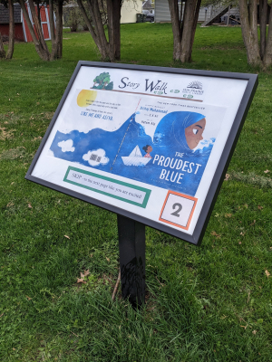 Photo of the Story Walk with the new book "The Proudest Blue." Grassy area, with a black post, and a panel attached to the post. In the panel is the Story Walk title/logo at the top, the book cover in the middle, an action to do at the bottom, and the number 2 in the lower right corner.