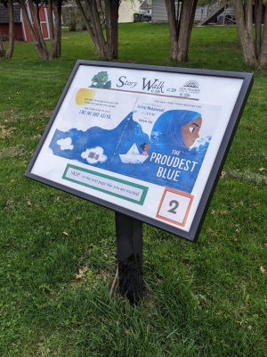 Story Walk - The Proudest Blue Photo of the Story Walk with the new book "The Proudest Blue." Grassy area, with a black post, and a panel attached to the post. In the panel is the Story Walk title/logo at the top, the book cover in the middle, an action to do at the bottom, and the number 2 in the lower right corner.