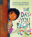 This image shows the book cover for the book "The Day You Begin" by Jacqueline Woodson. It shows a young girl stepping through a school classroom door and holding a book.